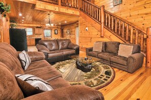 A log cabin interior with a living room featuring two brown leather sofas, a glass coffee table, and a wooden staircase. - Jennie's Cove | 6BR•Private Theater•Indoor Pool! (Sevierville)