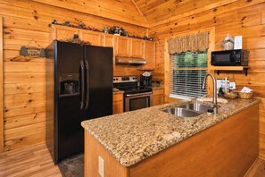 A cabin-style kitchen with wooden walls, a granite countertop, a stainless steel sink, a black refrigerator, a stove, and a microwave. - Charlie's Creek Retreat | Hot Tub • Game Loft! (Sevierville)