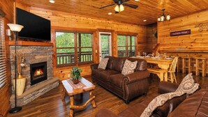 A log cabin interior with a fireplace, wooden floors, a sofa, a dining table, and a ceiling fan. - Savannah Splash | Hot Tub • Indoor Pool • Pets OK! (Sevierville)