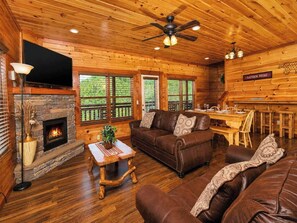 A log cabin interior with a fireplace, wooden floors, a sofa, a dining table, and a ceiling fan. - Savannah Splash | Hot Tub • Indoor Pool • Pets OK! (Sevierville)