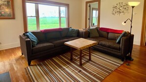 Een woonkamer met een hoekbank, een salontafel, een gestreept tapijt, een vloerlamp en een groot raam met uitzicht op een groen veld.