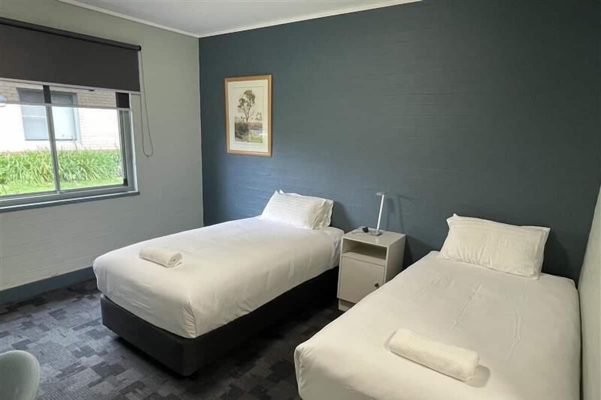 Habitación de hotel con dos camas, una ventana, una mesita de noche y un cuadro enmarcado en la pared.