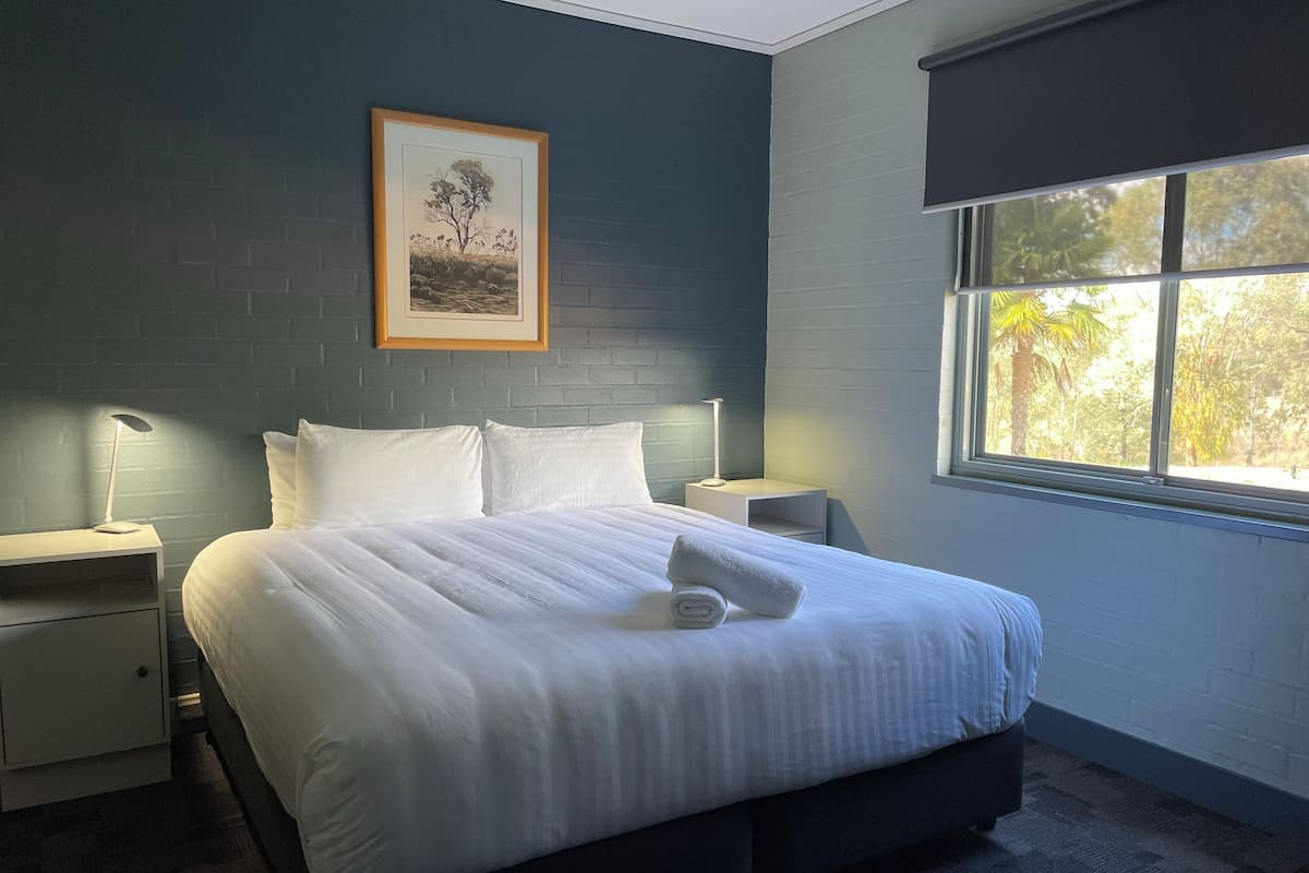 Una cama bien hecha con sábanas blancas, un cuadro en la pared y una ventana con vistas a árboles.