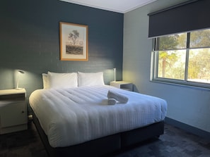 A neatly made bed with white linens, a framed picture on the wall, and a window with a view of trees. - Charles Sturt University - CPD Motel (Mitchell)