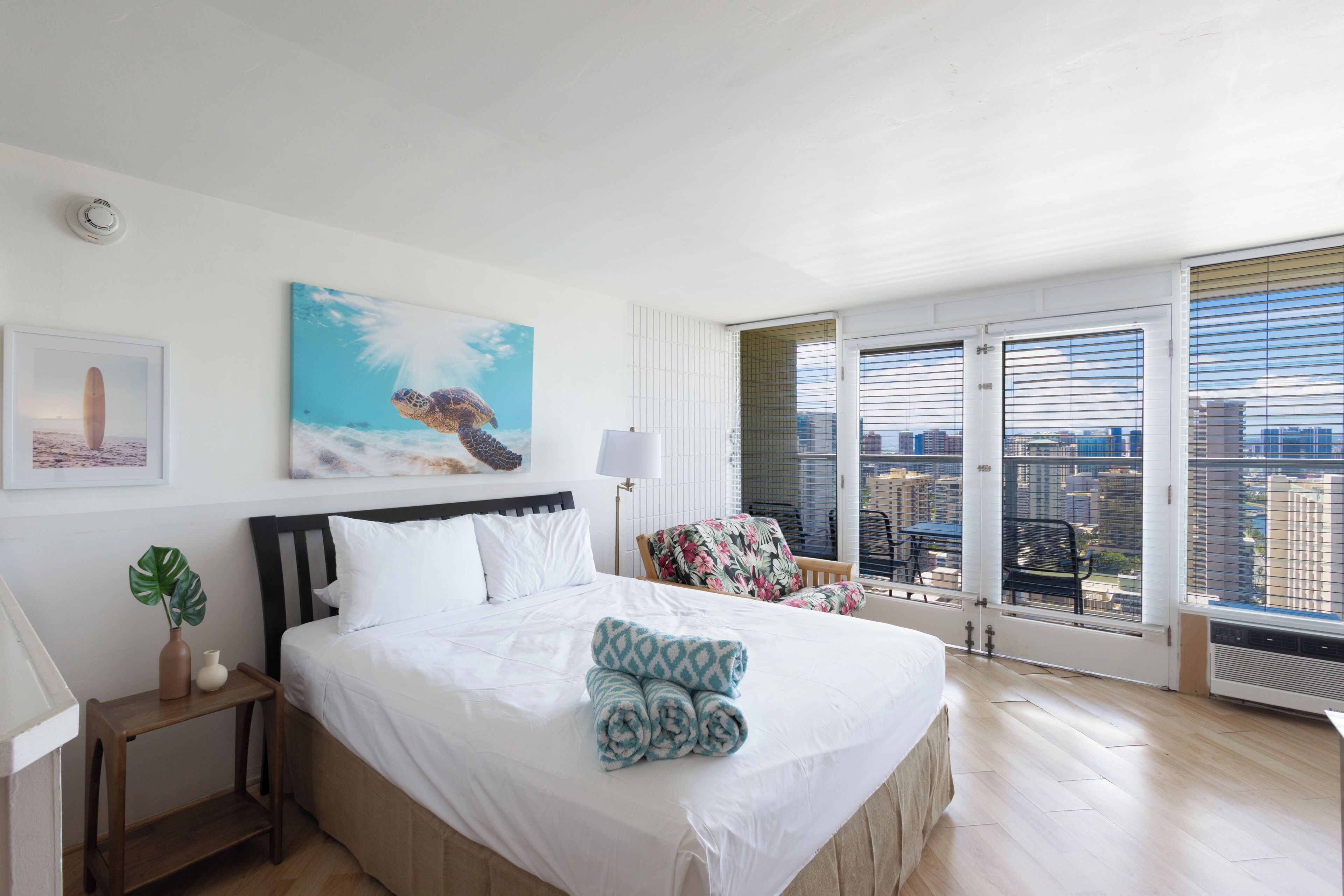A hotel room with a bed, a painting of a turtle, a chair, and a view of the city through large windows.