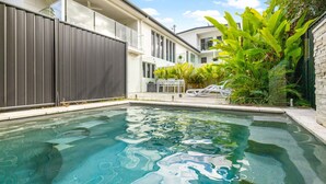Una piscina con agua cristalina, rodeada por una casa moderna con balcón y abundante vegetación verde.