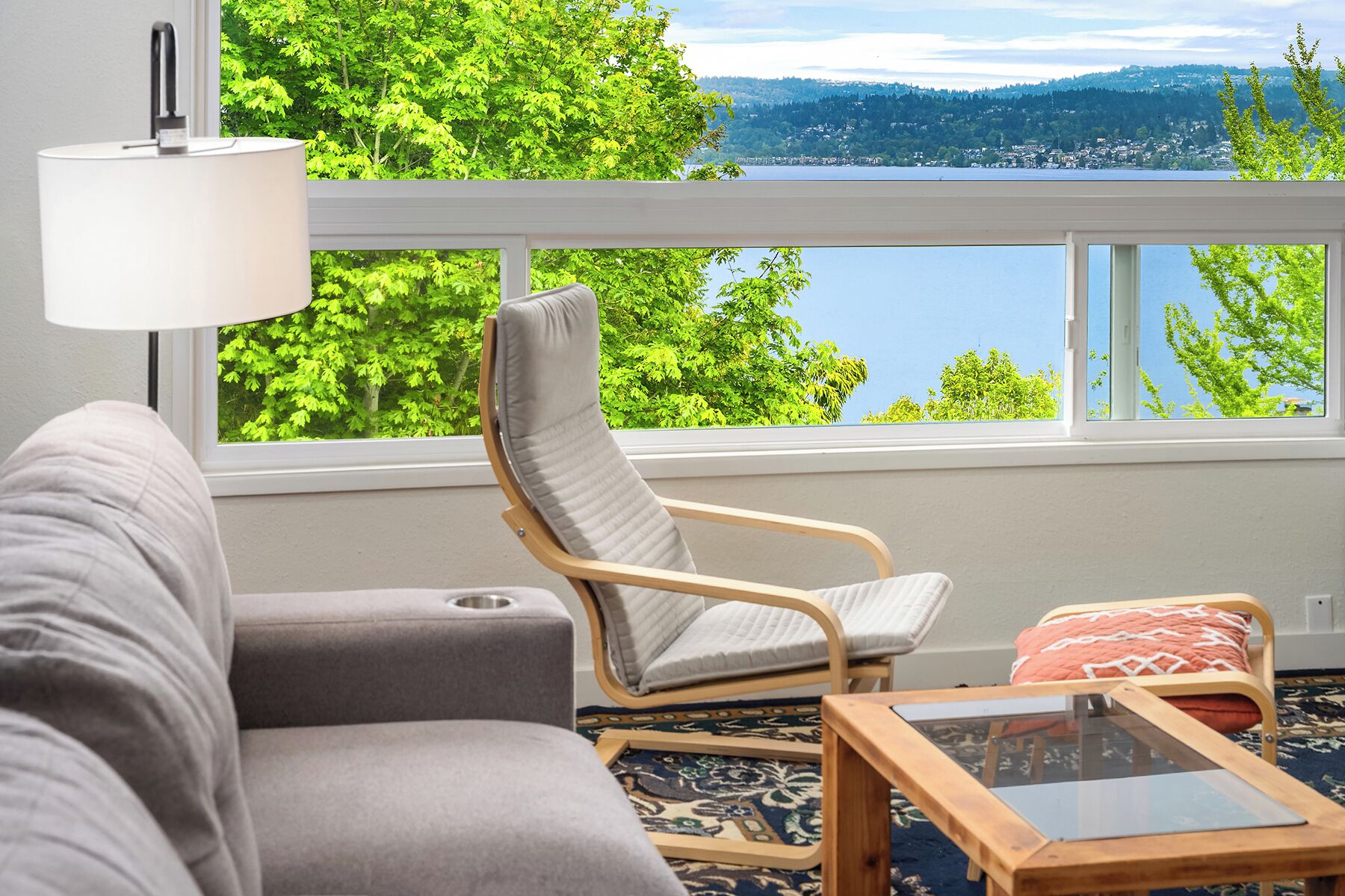 A living room with a view of a lake and mountains, a sofa, a chair, and a small table.
