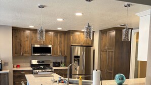 A modern kitchen with wooden cabinets, stainless steel appliances, a central island with bar stools, pendant lighting, and a large window.