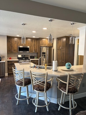 A modern kitchen with wooden cabinets, stainless steel appliances, a central island with bar stools, pendant lighting, and a large window.