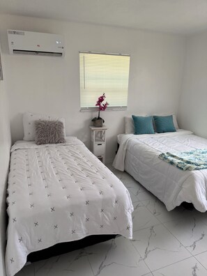 Zwei Einzelbetten mit weißer Bettwäsche, ein kleiner Tisch mit einem Blumentopf, ein Fenster mit Jalousien und eine Klimaanlage an der Wand.