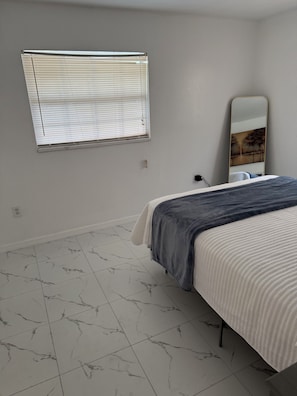 A bedroom with a bed, a window with blinds, a mirror, and a marble-patterned floor.