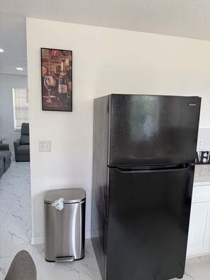 A black refrigerator, a stainless steel trash bin, a gray couch, and a framed picture on the wall.