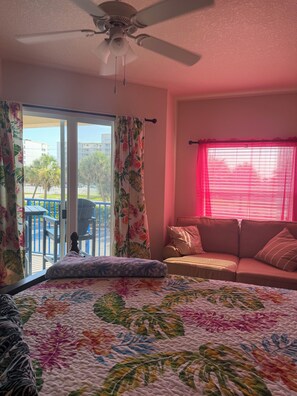 Ein Schlafzimmer mit Bett, Deckenventilator, Balkon mit Stuhl und Fenster mit Vorhängen.