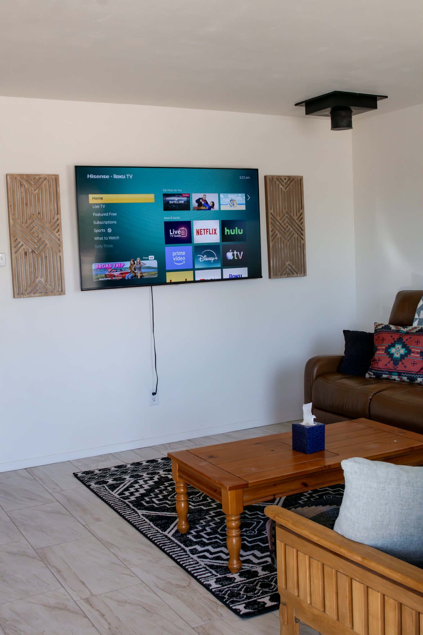 Ein Wohnzimmer mit an der Wand befestigtem Fernseher, der verschiedene Streaming-Dienste anzeigt, einem braunen Ledersofa, einem Holztisch und einem gemusterten Teppich.