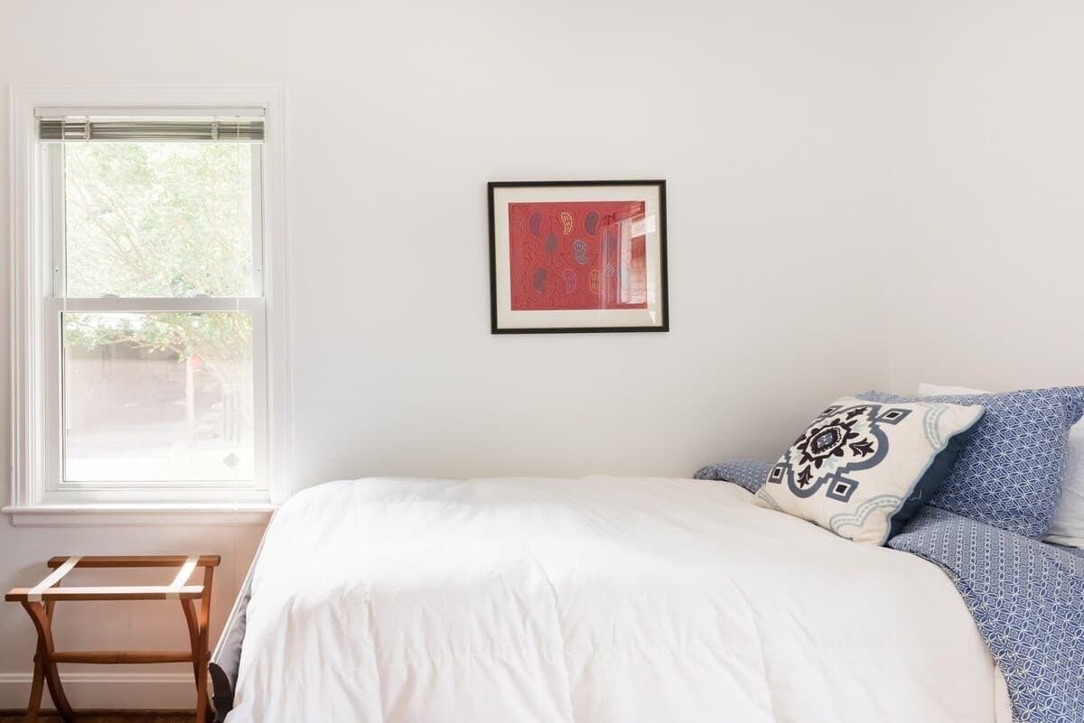 Un dormitorio con cama, ventana con persianas, un cuadro enmarcado en la pared y una pequeña mesa de madera.