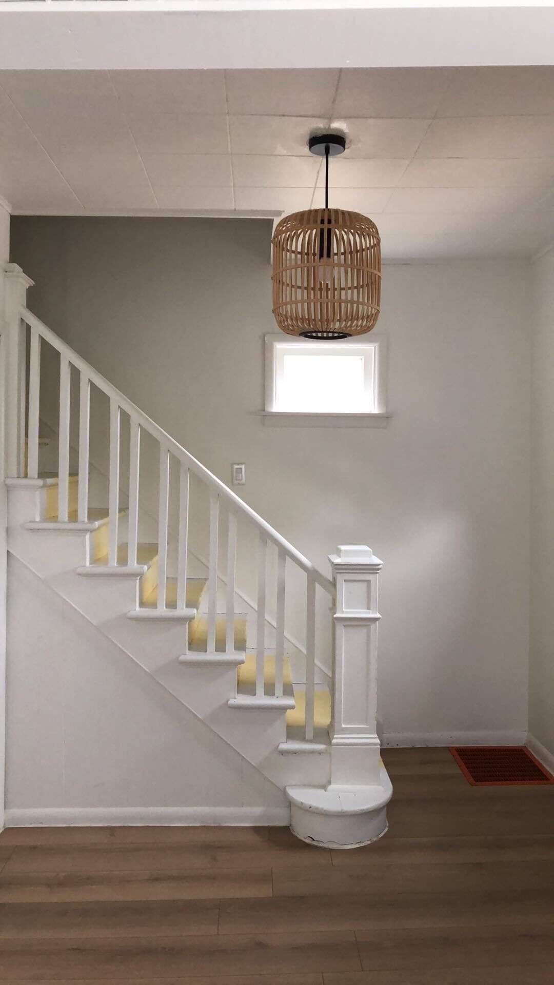 Eine Treppe mit weißen Geländern und gelben Stufen, eine gewebte Pendelleuchte und ein kleines Fenster.