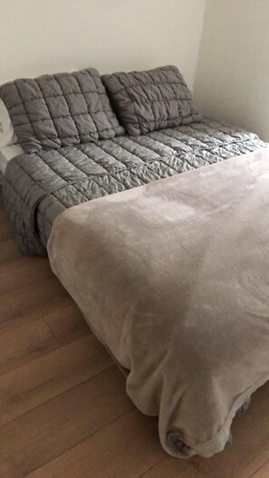 A bed with a grey quilt and pillows, a wooden floor, and a plain wall. - The Downtown Hamilton House (Shédiac)