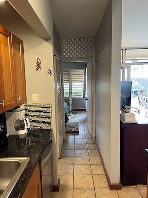 A narrow hallway with a kitchen on one side, a dining area with a table and chairs on the other, and a view of a balcony with a railing. - Perfect 1-bedroom condo on Molokai's West End for your getaway (Maunaloa)