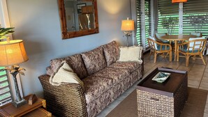 A living room with a sofa, coffee table, wicker side tables, a lamp, a mirror, and a dining area with chairs and a table.