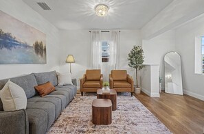 Ein Wohnzimmer mit grauem Sofa, zwei braunen Sesseln, einem Couchtisch, einem Gemälde, einer Lampe, einer Pflanze und einem Spiegel.