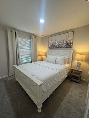 A bedroom with a white bed, two bedside lamps, a painting above the bed, and a window with curtains. - Tranquil 3-bedroom house in charming Jacksonville Minutes to San Marco& Downtown (Jacksonville)