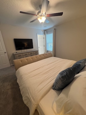 Ein Schlafzimmer mit Bett, Deckenventilator, Fernseher an der Wand, Kommode und einer Tür.