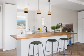 A modern kitchen with a wooden island, bar stools, pendant lights, a view of the city, and a painting on the wall. - Le Panoramique Niçois avec Terrasse - Welkeys (Nice)
