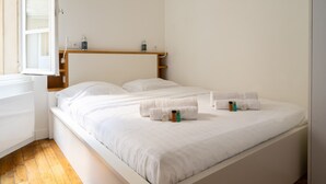 Ein ordentlich bezogenes Bett mit weißen Leinen und Handtüchern, ein Holz-Kopfteil und ein kleines Regal mit Pflegeprodukten.