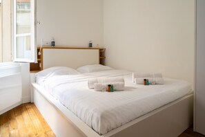 Ein ordentlich bezogenes Bett mit weißen Leinen und Handtüchern, ein Holz-Kopfteil und ein kleines Regal mit Pflegeprodukten.
