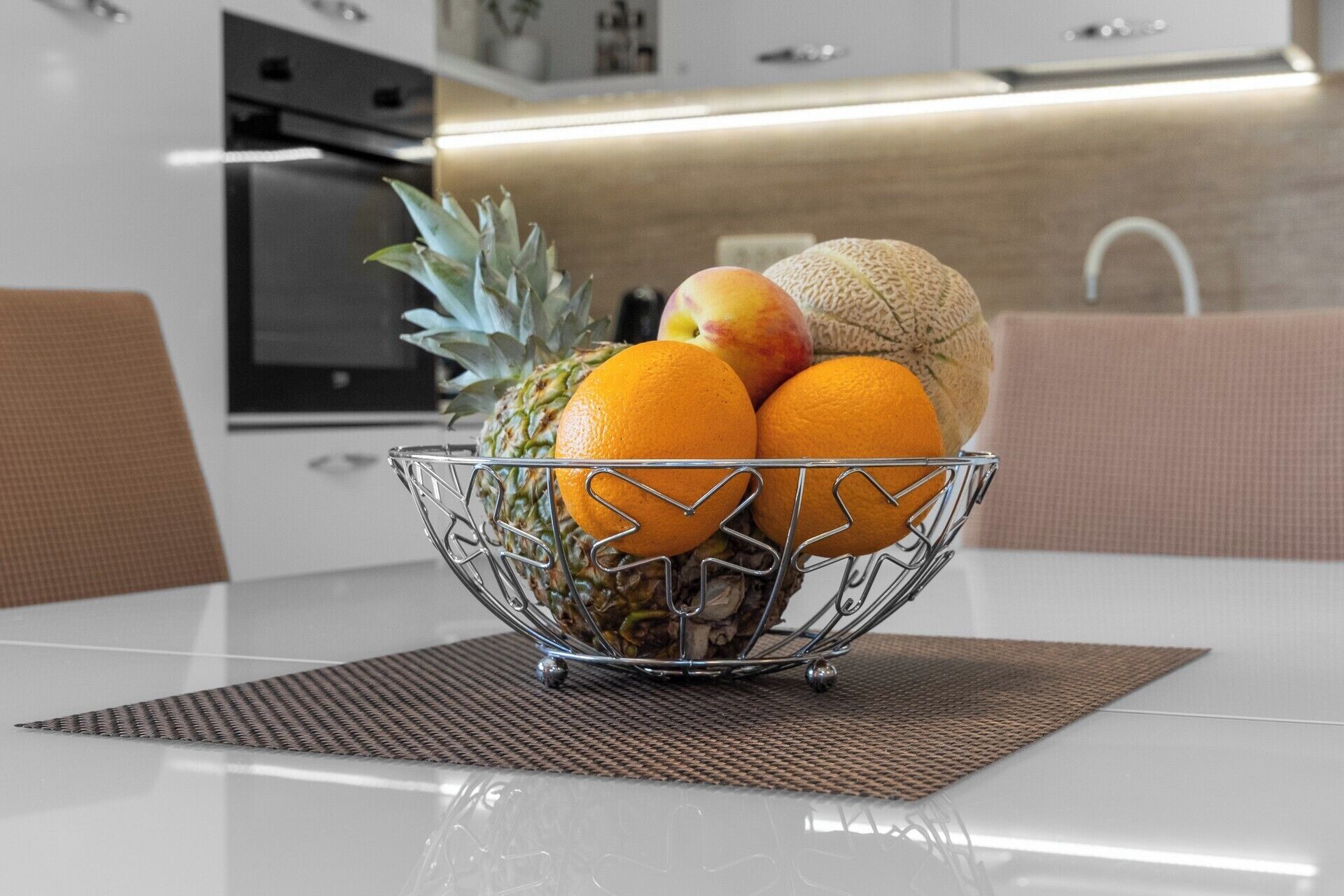 A fruit bowl on a table with a woven placemat, a kitchen in the background, and a modern design.