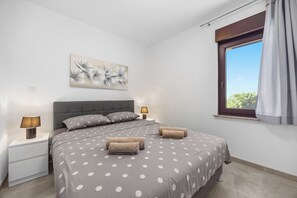 Ein Schlafzimmer mit einem Bett, zwei Nachttischlampen, einem Fenster mit Vorhängen und einem an der Wand hängenden Kunstwerk.