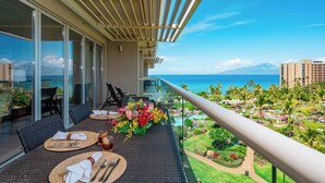 A balcony with a dining table set for two, overlooking a resort area with palm trees and a view of the ocean. - 🌊 Hokulani 746 Penthouse with Unobstructed Ocean Views - Special Intro Rates! (Lahaina)