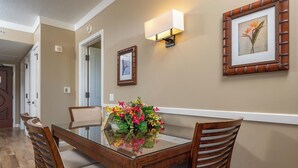 A dining area with a glass-top table, wooden chairs, a floral centerpiece, wall-mounted light fixtures, and framed artwork. - 🌊 Hokulani 746 Penthouse with Unobstructed Ocean Views - Special Intro Rates! (Lahaina)