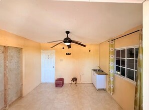 Habitació amb ventilador de sostre, una porta blanca, una cuineta petita i una finestra amb cortines florals.