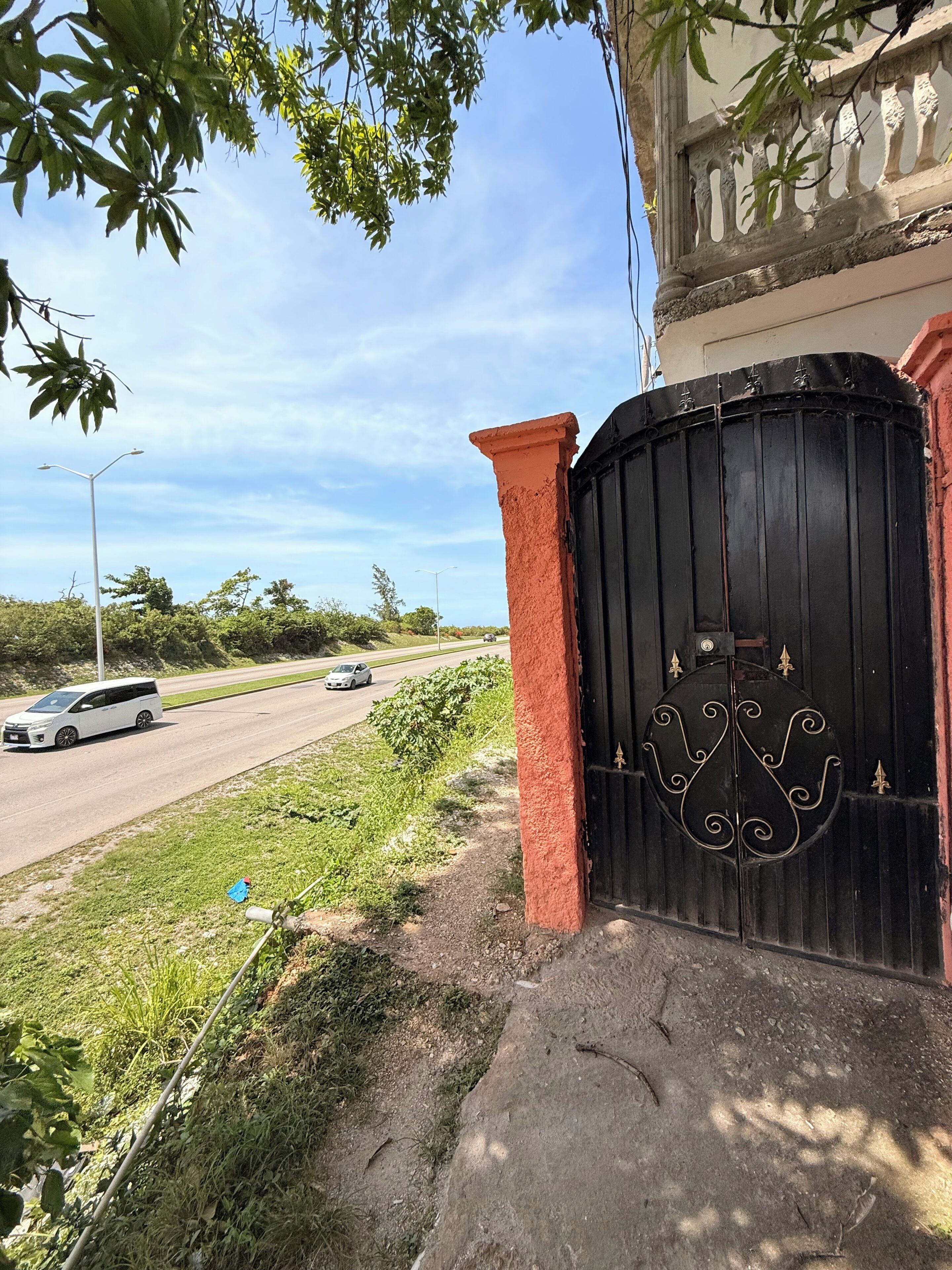 A gated entrance with a black gate, a red pillar, and a street with passing vehicles.