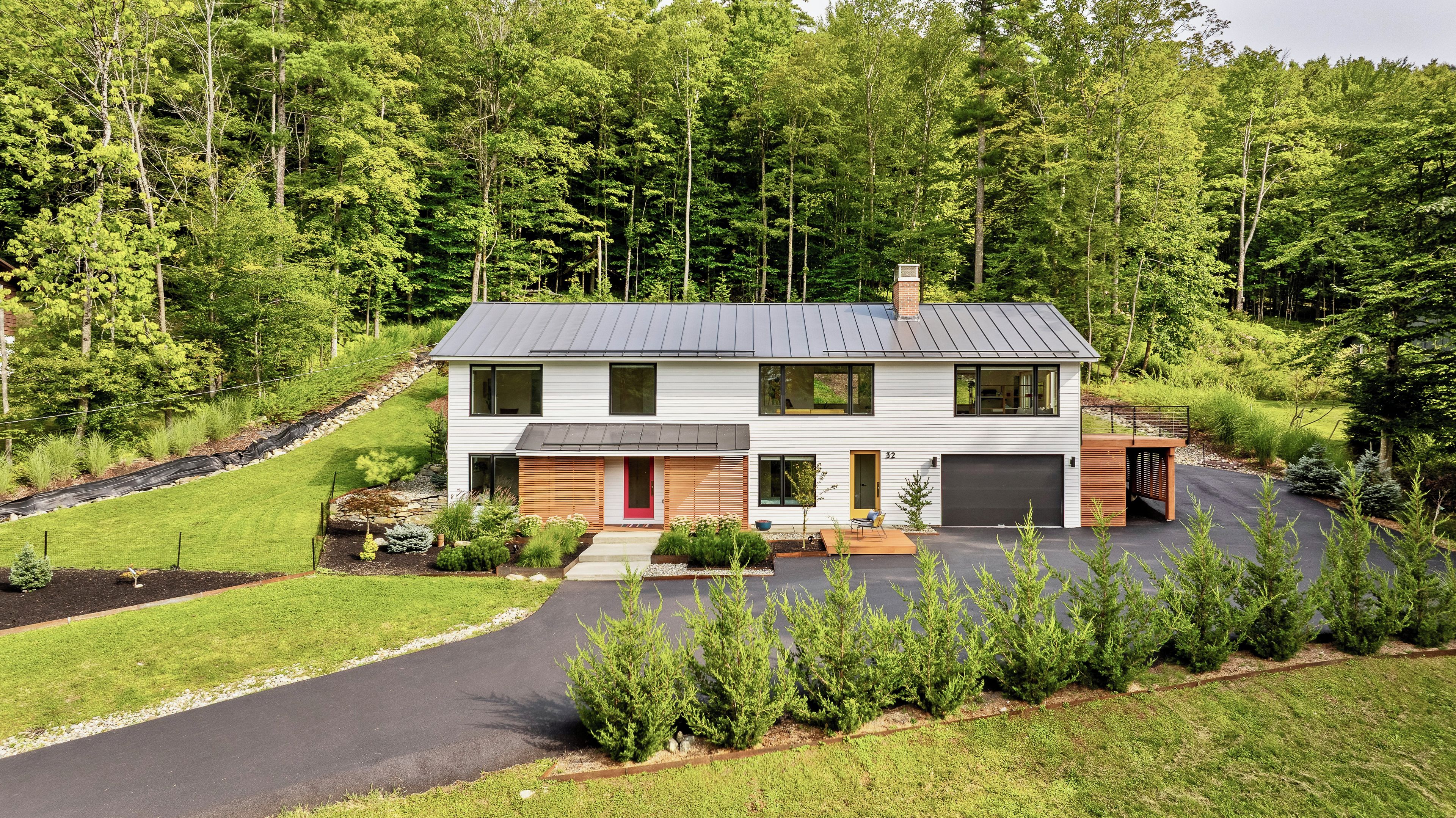 Une maison moderne à deux étages avec un garage, entourée d’une pelouse bien entretenue et d’une forêt dense.