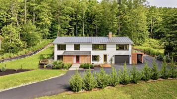 Une maison moderne à deux étages avec un garage, entourée d’une pelouse bien entretenue et d’une forêt dense.
