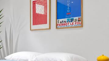 A modern bedroom with a white bed, a small bedside table with a book, and two framed posters on the wall.
