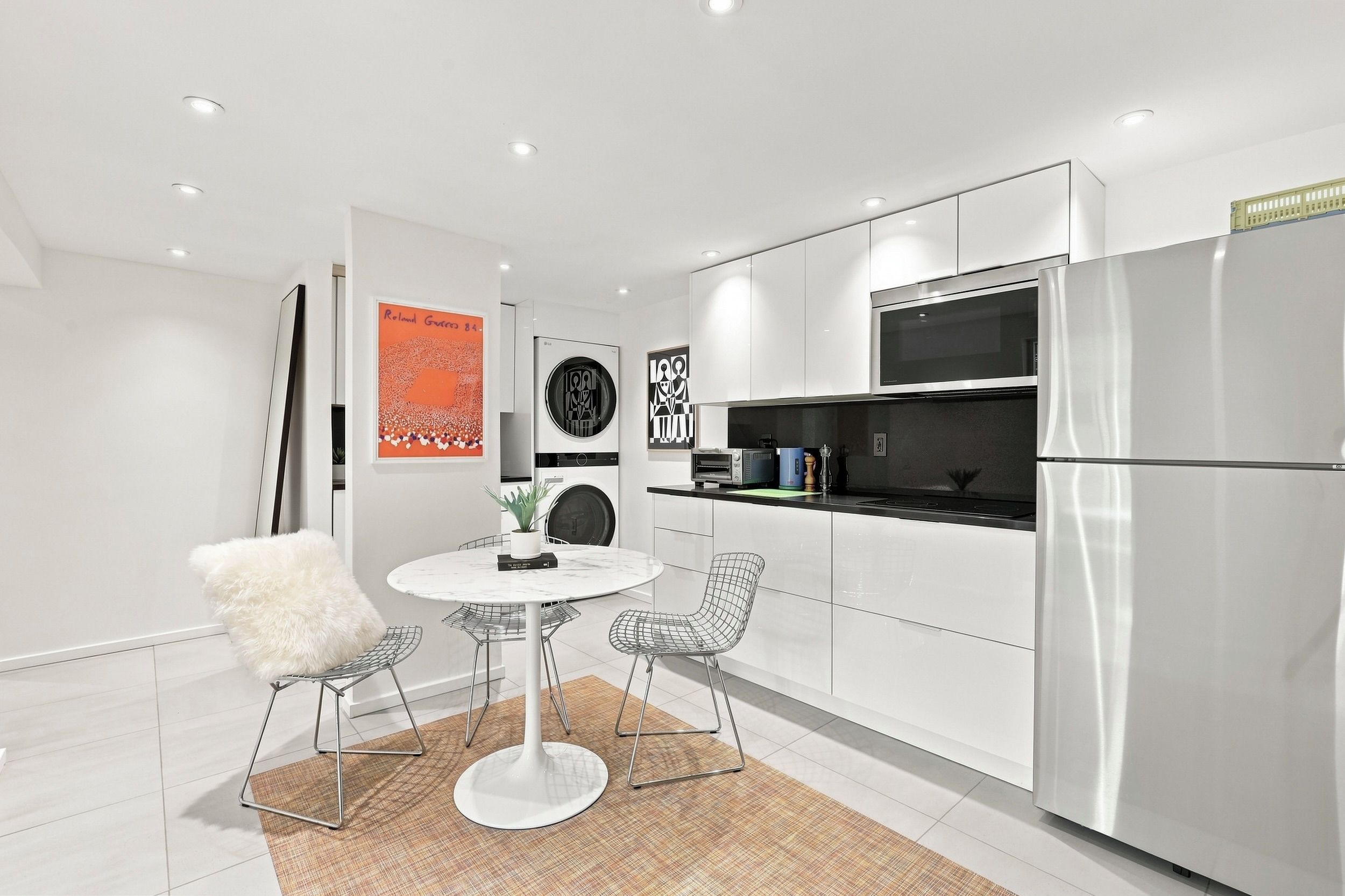 Une cuisine moderne avec des meubles blancs, un réfrigérateur en acier inoxydable, un four à micro-ondes et un four encastré. On y trouve une table de salle à manger ronde, accompagnée de deux chaises et d’un fauteuil blanc moelleux.