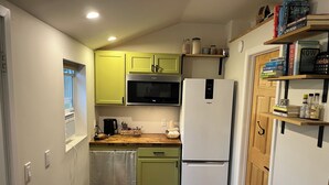 A compact kitchen with a microwave, refrigerator, and wooden countertop. There are books and jars on shelves, and a bench is visible.