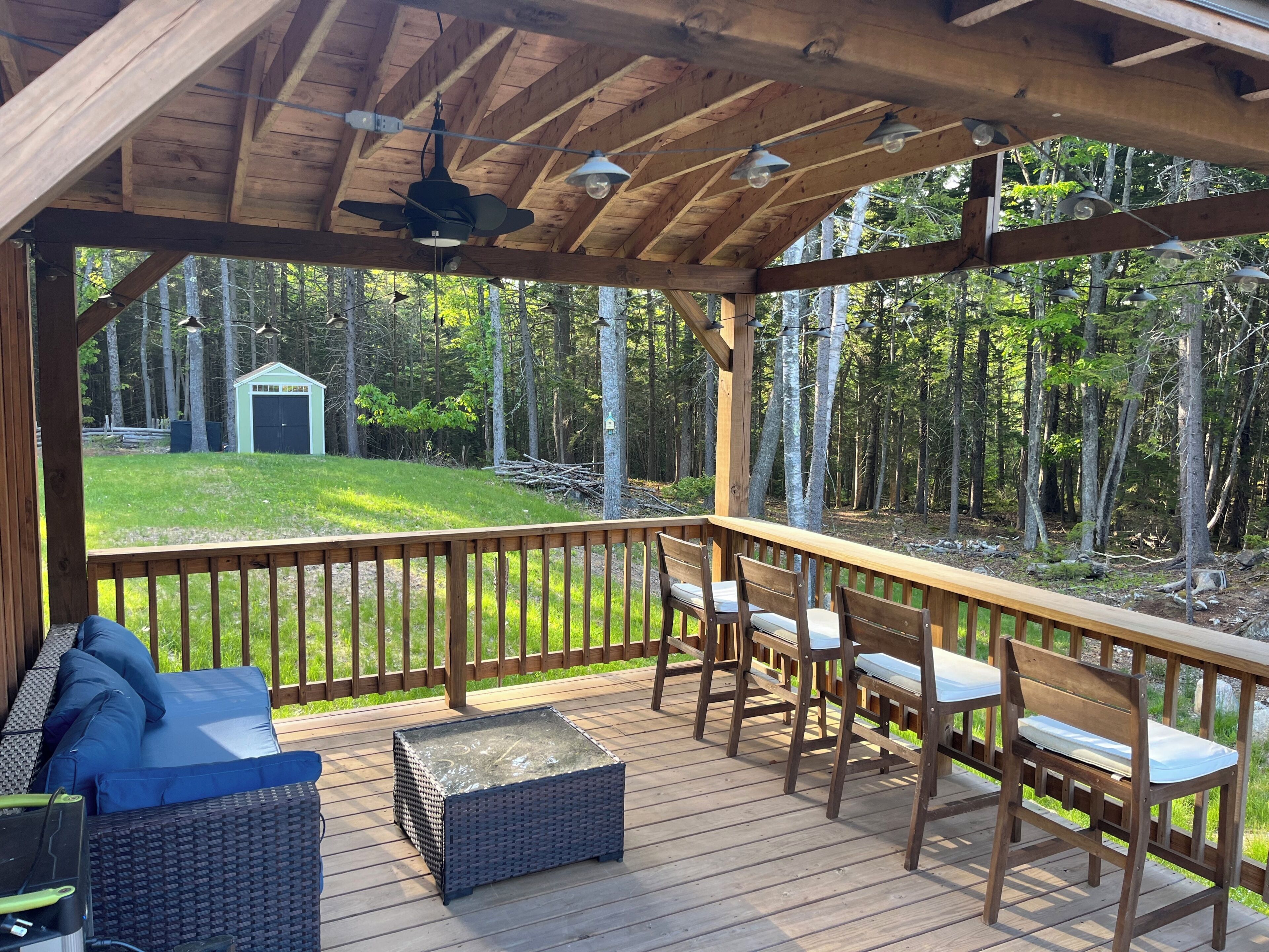 A covered deck with wooden furniture, a glass-top table, and a view of a forested yard.