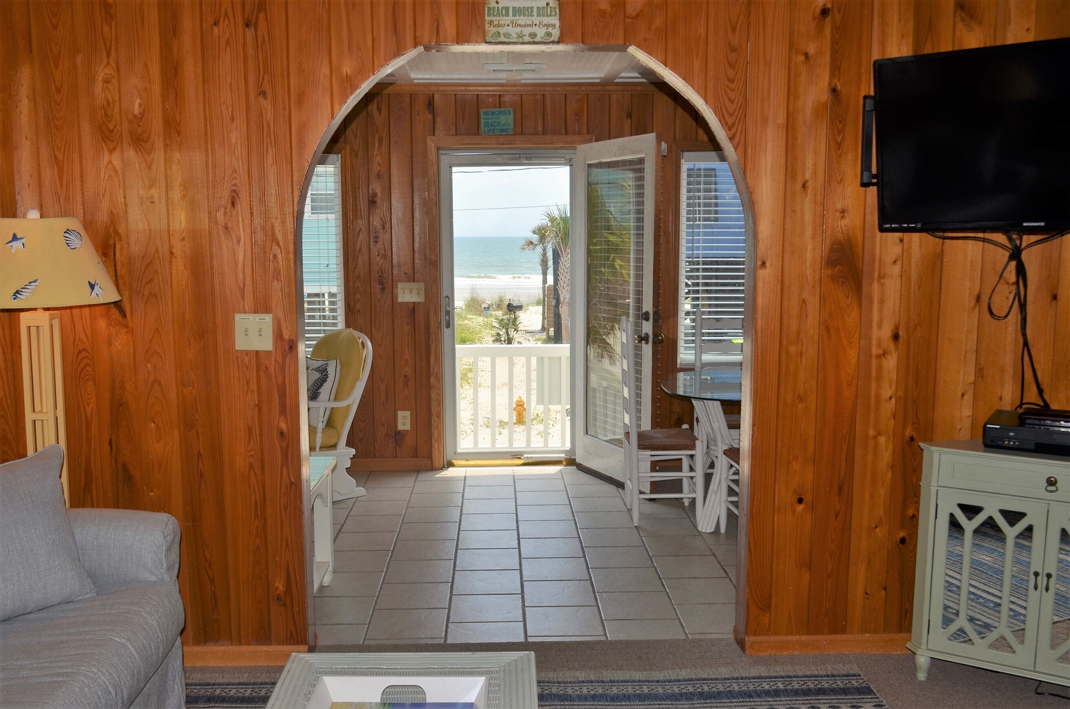 A wooden interior with a view to the beach, a television, and a chair by the door.