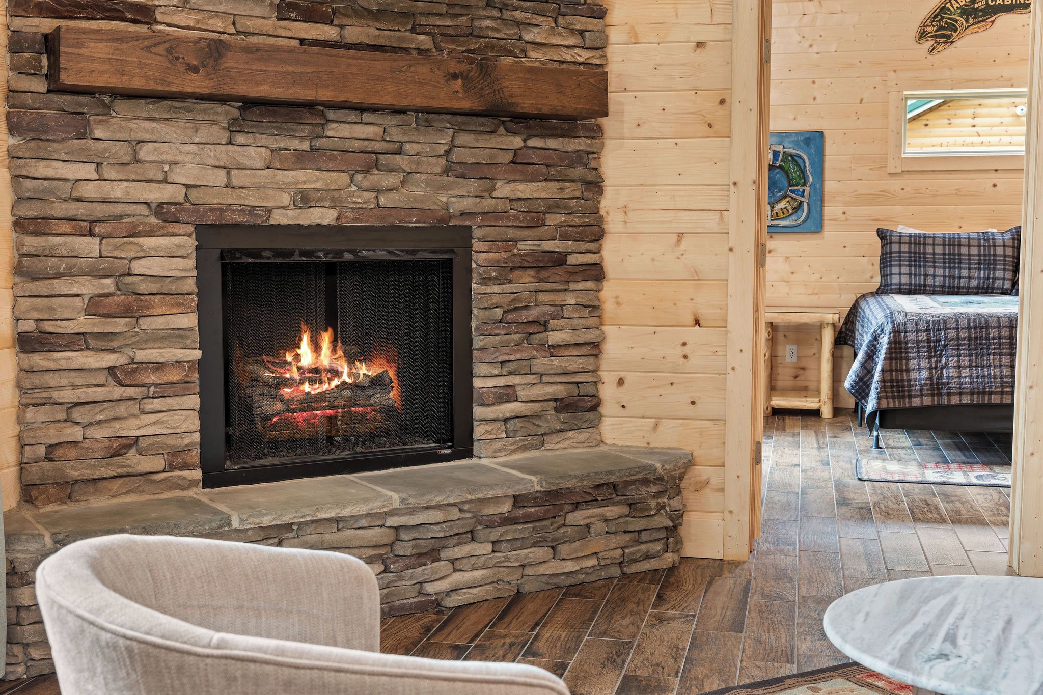 A stone fireplace with a fire, a wooden wall, a bed with a plaid bedspread, and a chair.