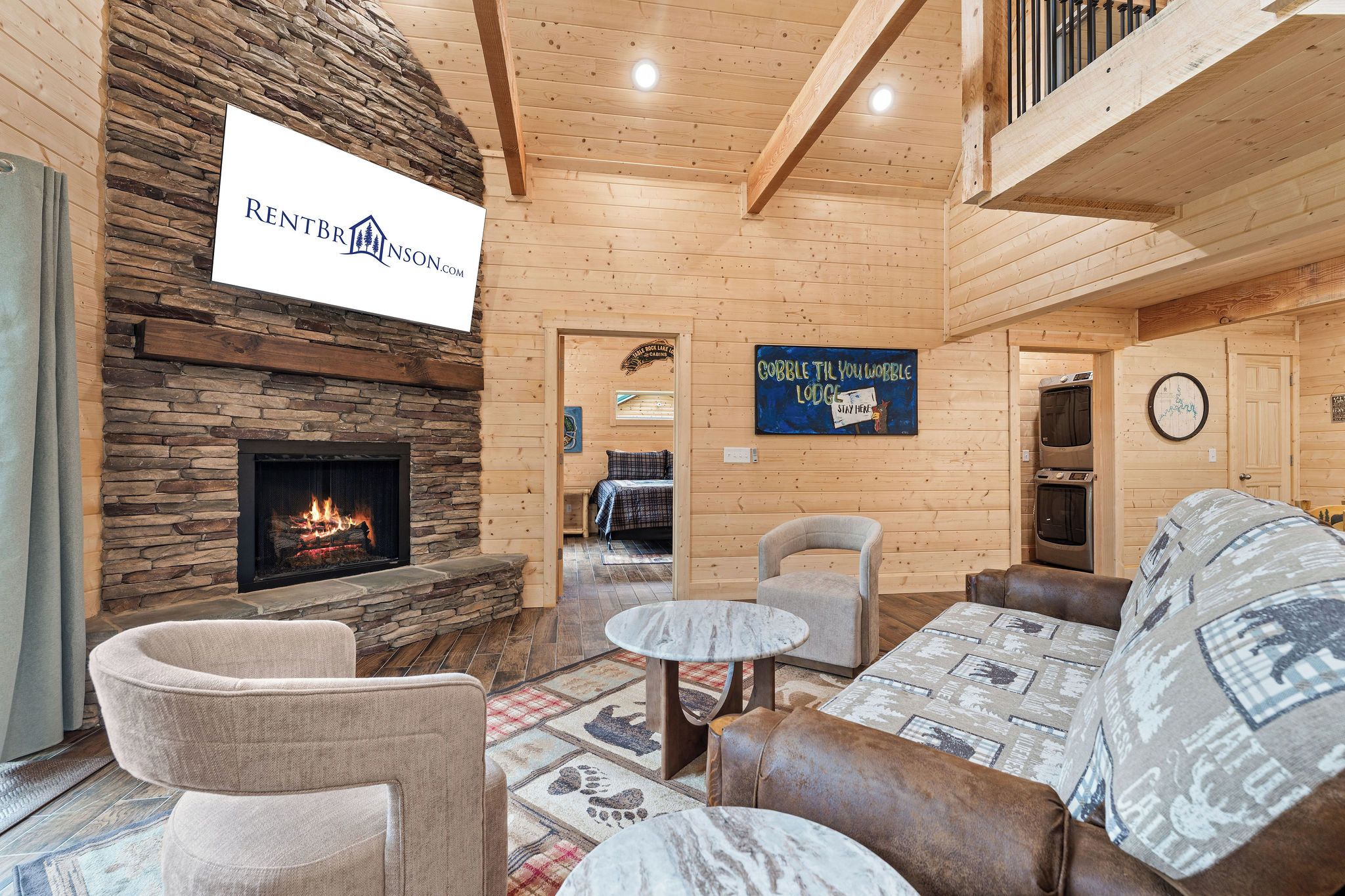 A cozy interior with a stone fireplace, wooden walls, a sign for RentBrinson.com, and a sign that says 'Cobblestone Till You Wobble Lodge Stay Here'.