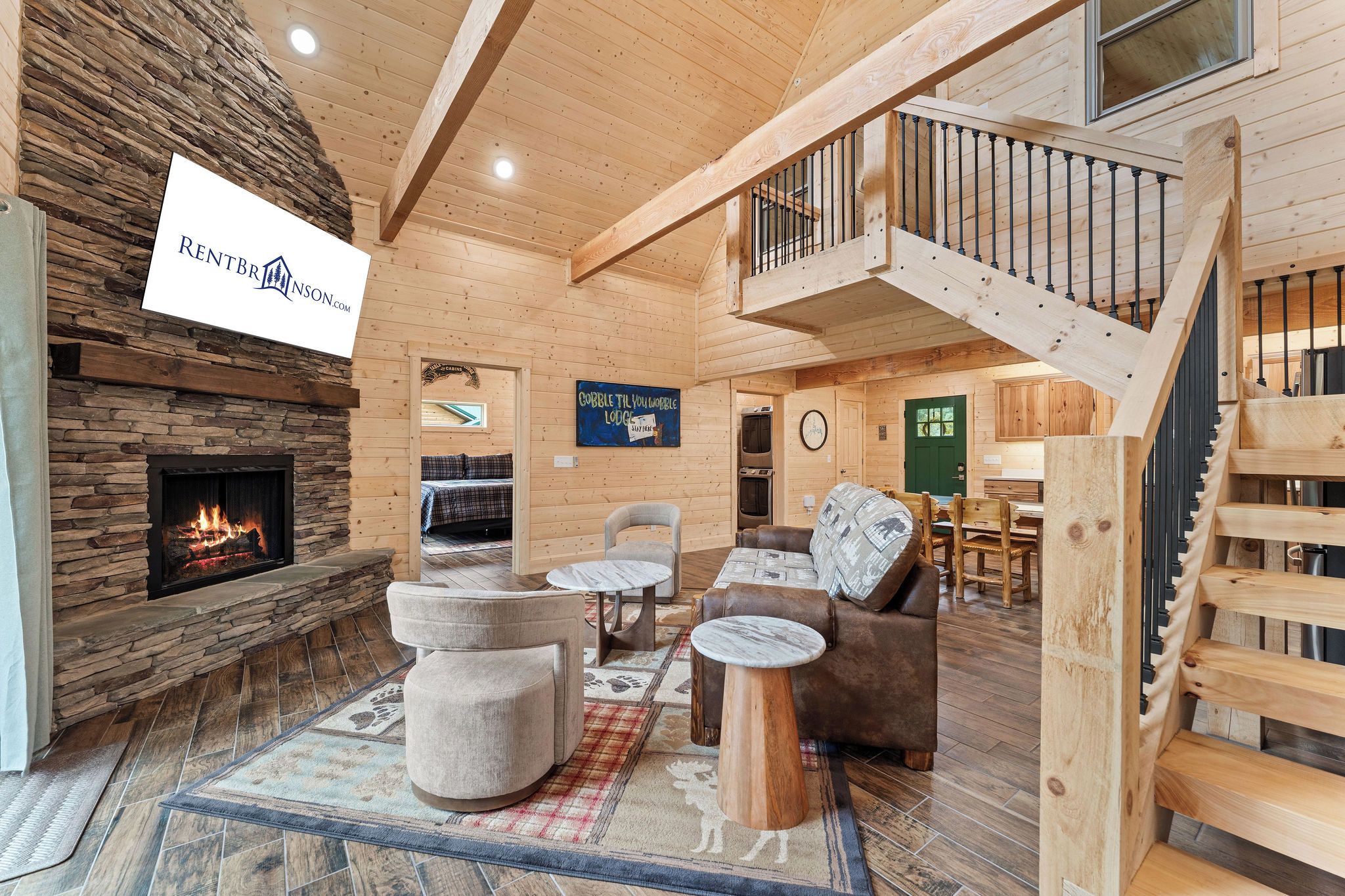 A cozy lodge interior with a stone fireplace, wooden furniture, and a staircase leading to an upper level.