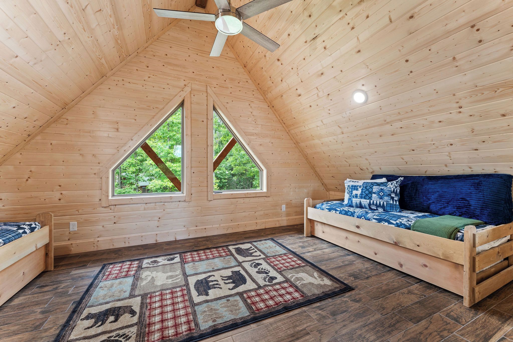 A cozy attic bedroom with a sloped ceiling, two windows, a bed, a sofa, and a patterned rug.