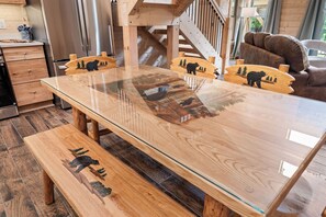 A wooden dining table with glass top and bear carvings, set in a room with a staircase and a sofa.