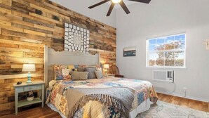 Ein Schlafzimmer mit Holzwand, einem Bett mit Blumenmuster auf der Tagesdecke, einem Nachttisch mit Lampe und einem Fenster mit Blick.