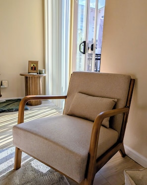 Ein modernes Wohnzimmer mit einem beigefarbenen Sessel, einem kleinen Holztisch und Blick auf ein Gebäude durch große Fenster.
