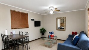 Uma sala de estar com sofá, mesa de jantar com cadeiras, televisão, ventilador de teto e espelho.